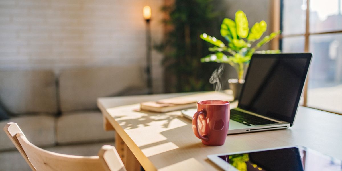 Laptop and red mug on a wooden desk with a cozy living room in the background.